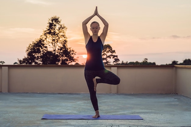 Giovane donna atletica che pratica yoga sul tetto al tramonto, silhouette.