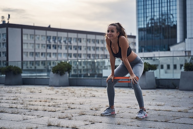 Giovane donna atletica che sorride mentre fa esercizi di squat con gomma sportiva nella città urbana