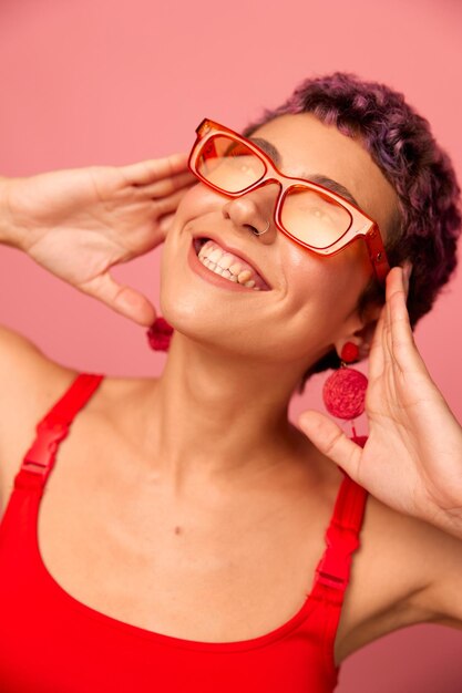 Giovane donna atletica con un taglio di capelli corto e capelli viola in un top rosso e leggings da yoga rosa in occhiali da sole con una figura atletica sorride e balla su uno sfondo rosa