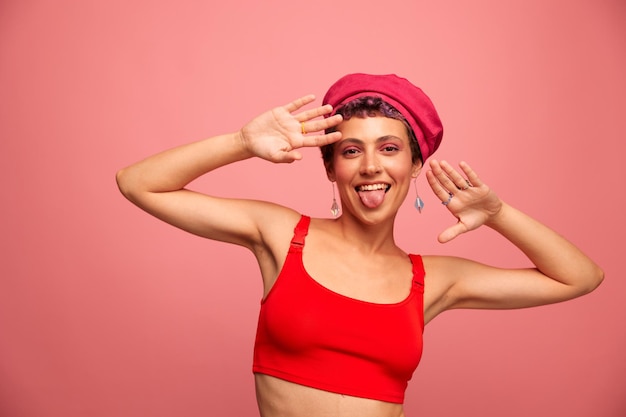 Giovane donna atletica con un taglio di capelli corto e capelli viola in un top rosso e un cappello rosa con una figura atletica sorride e fa smorfie guardando la telecamera su uno sfondo rosa