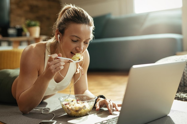 Giovane donna atletica sdraiata sul pavimento e mangiare cibo sano mentre naviga in rete sul computer portatile.