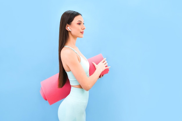 Giovane donna atletica sorridente in abiti sportivi in posa contro una parete blu dello studio. Il concetto di allenamento motivazionale sportivo. Copia spazio. Tieni un tappetino da yoga