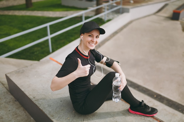 Giovane donna atletica sorridente in uniforme nera, berretto con le cuffie che ascolta la musica, tenendo la bottiglia con acqua, seduta sull'allenamento, mostrando il pollice nel parco cittadino all'aperto