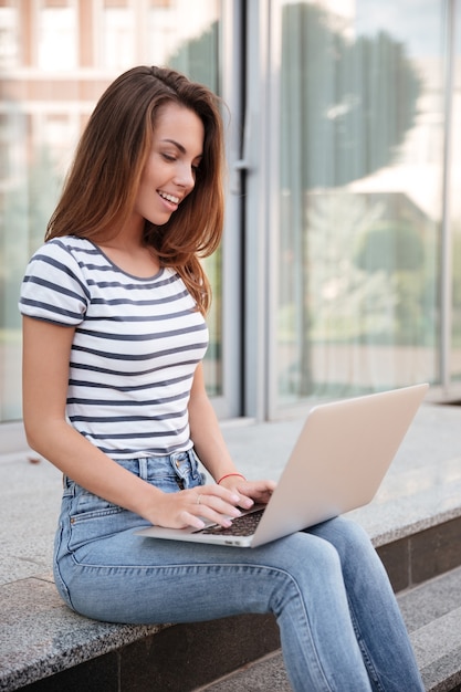 Giovane donna attraente allegra che sorride e che usa il computer portatile all'aperto