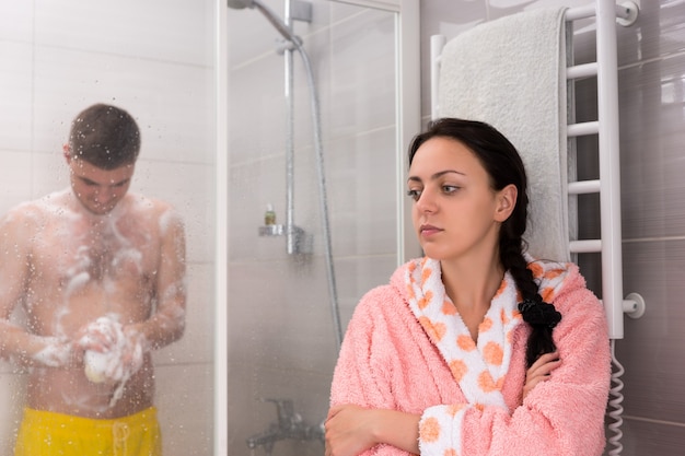 Giovane donna attraente che aspetta il suo ragazzo mentre si strofina una spugna da bagno in schiuma nella cabina doccia con porte in vetro trasparente in bagno