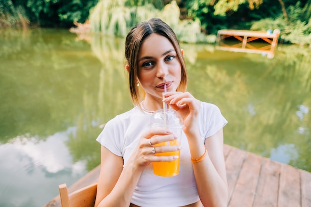 Giovane donna attraente che beve limonata arancione, riposante su un molo sul lago all'aperto.