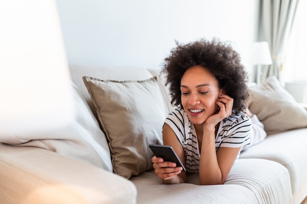 Giovane donna attraente che esamina il suo Smart Phone e che sorride mentre sedendosi sul sofà a casa.