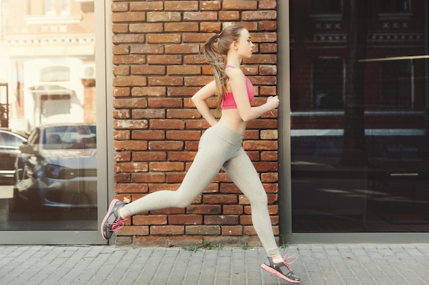 Giovane donna attraente che fa jogging in città vicino a un edificio in mattoni con finestre a specchio, copia spazio, vista laterale