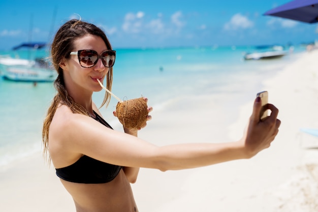 Giovane donna attraente che fa un selfie mentre bevendo il cocktail di pinacolada su una bella spiaggia soleggiata.