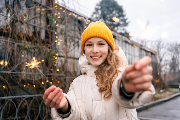 Giovane donna attraente che guarda la telecamera e sorride mentre tiene in mano due stelle filanti sullo sfondo di una serra con ghirlande Il concetto di vacanze di Capodanno
