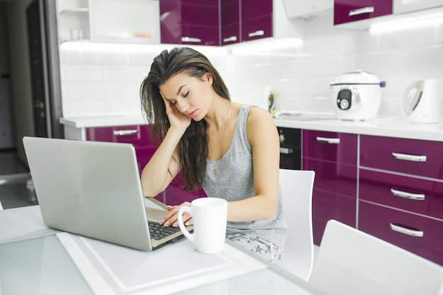 Giovane donna attraente che lavora al pc in cucina. Lavoro femminile al computer di mattina. Libero professionista al computer portatile al coperto. Imprenditrice che lavora a casa.