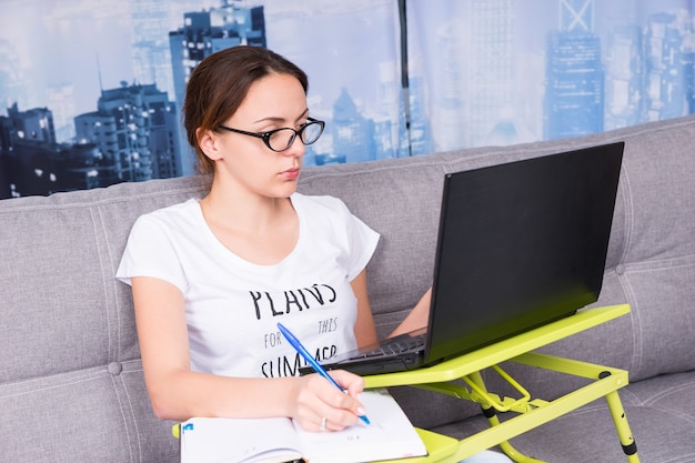 Giovane donna attraente con gli occhiali che lavora su un laptop facendo i suoi affari da casa con in mano una penna mentre legge le informazioni sullo schermo