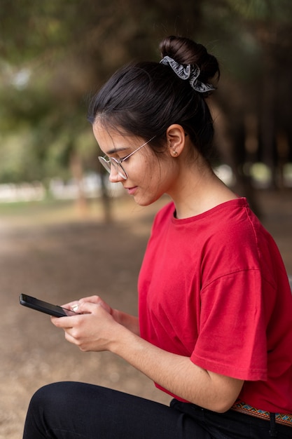 Giovane donna attraente con gli occhiali seduto su una panchina e usando il suo telefono in un parco, con una camicia rossa. Siviglia, Spagna