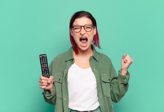 Giovane donna attraente con i capelli rossi che grida in modo aggressivo con un'espressione arrabbiata o con i pugni chiusi per celebrare il successo e tenere in mano un telecomando della tv