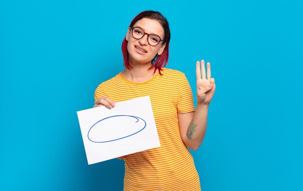 Giovane donna attraente con i capelli rossi che sorride e sembra amichevole, mostrando il numero tre o terzo con la mano in avanti, conto alla rovescia