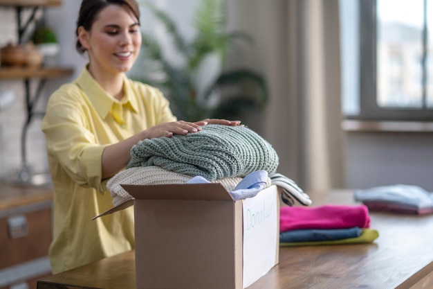 Giovane donna attraente concentrata soddisfatta sorridente che mette due pullover piegati della maglieria nella scatola di cartone