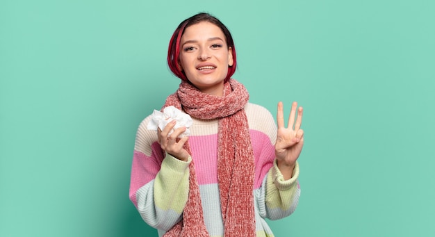 Giovane donna attraente dai capelli rossi che sorride e sembra amichevole, mostrando il numero tre o il terzo con la mano in avanti, contando il concetto di influenza