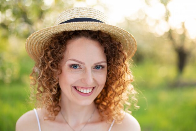 Giovane donna attraente del ritratto con capelli ricci in un cappello di vimini alla moda in un giardino verde