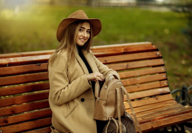 Giovane donna attraente in abiti autunnali si siede su una panchina nel parco cittadino. Donna vestita con cappotto elegante e cappello di feltro. Tempo d'autunno
