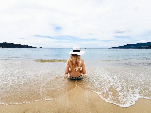 Giovane donna attraente in un cappello sulla riva del mare blu che si siede indietro con una noce di cocco, Tailandia