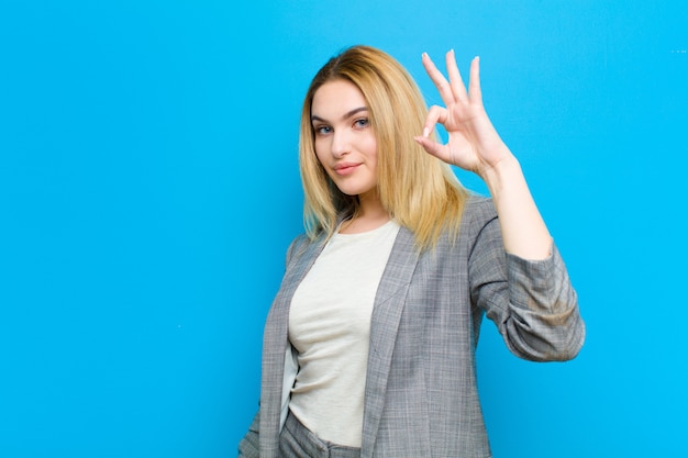 Giovane donna bella bionda sentirsi felice, rilassato e soddisfatto, mostrando l'approvazione con gesto ok, sorridendo contro la parete piatta