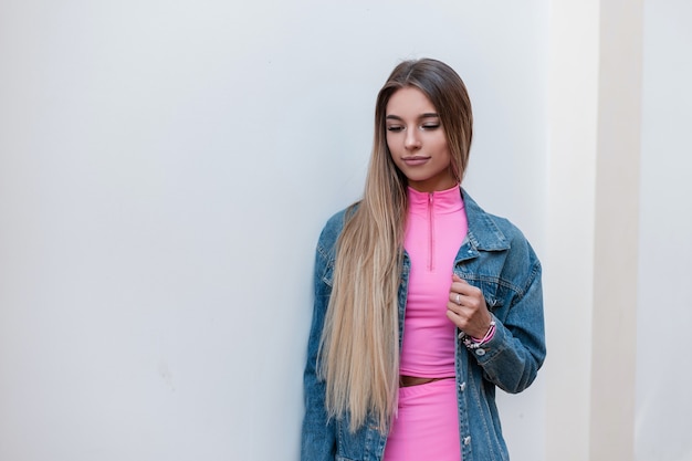 Giovane donna bionda abbastanza affascinante in un vestito sportivo estivo rosa alla moda in una giacca di jeans blu alla moda che gode di un periodo di riposo in città. Modello di moda bella ragazza carina si trova vicino al muro bianco.