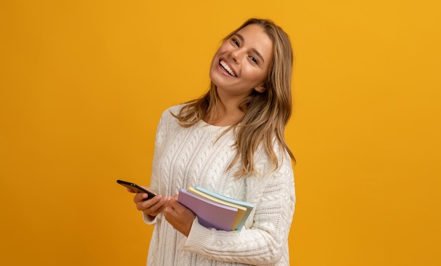 Giovane donna bionda abbastanza sorridente che tiene in mano utilizzando lo smartphone in posa con libri su sfondo giallo studio isolato felice educazione che indossa un maglione bianco