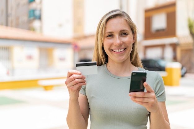 Giovane donna bionda all'aperto che acquista con il cellulare con una carta di credito
