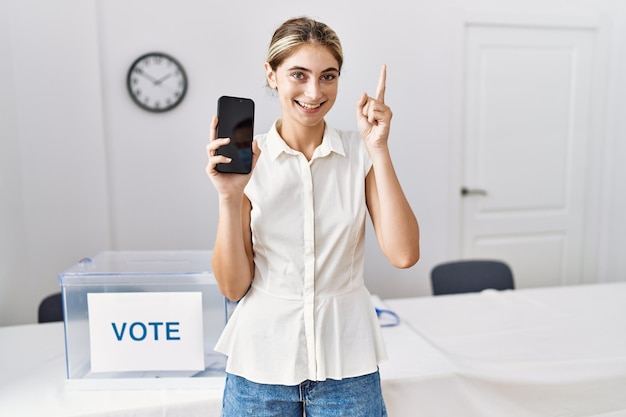 Giovane donna bionda alle elezioni politiche che tiene smartphone sorridente con un'idea o una domanda che punta il dito con la faccia felice numero uno