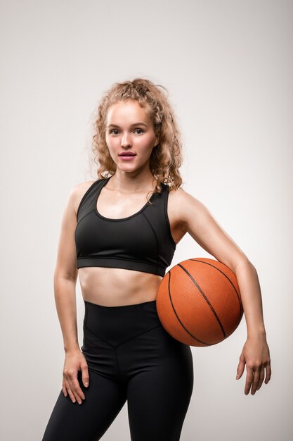 Giovane donna bionda attiva in abbigliamento sportivo che tiene la palla tra la vita e il braccio mentre si trova su gray