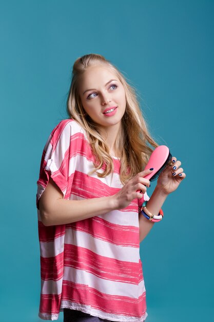 Giovane donna bionda attraente che si spazzola i capelli con un pettine rosa su sfondo blu