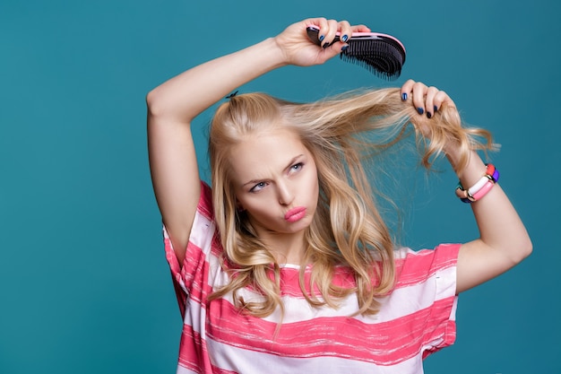 Giovane donna bionda attraente che si spazzola i capelli con un pettine rosa su sfondo blu