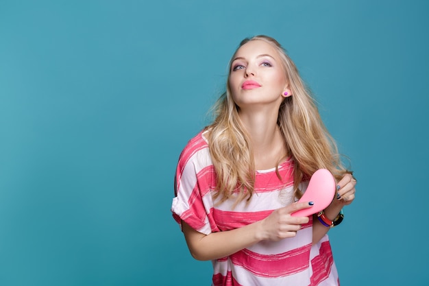 Giovane donna bionda attraente che si spazzola i capelli con un pettine rosa su sfondo blu