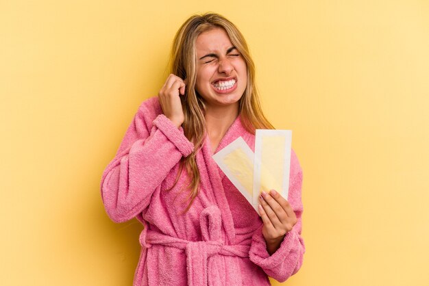 Giovane donna bionda caucasica che indossa accappatoio che tiene fasce depilatorie isolate su sfondo giallo che copre le orecchie con le mani.