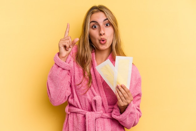 Giovane donna bionda caucasica che indossa accappatoio che tiene fasce depilatorie isolate su sfondo giallo con qualche grande idea, concetto di creatività.