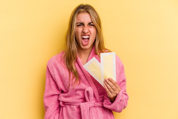 Giovane donna bionda caucasica che indossa accappatoio che tiene fasce depilatorie isolate su sfondo giallo urlando molto arrabbiato e aggressivo.