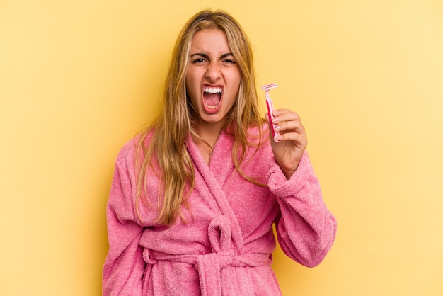 Giovane donna bionda caucasica che indossa accappatoio che tiene le lame isolate su sfondo giallo urlando molto arrabbiato e aggressivo.