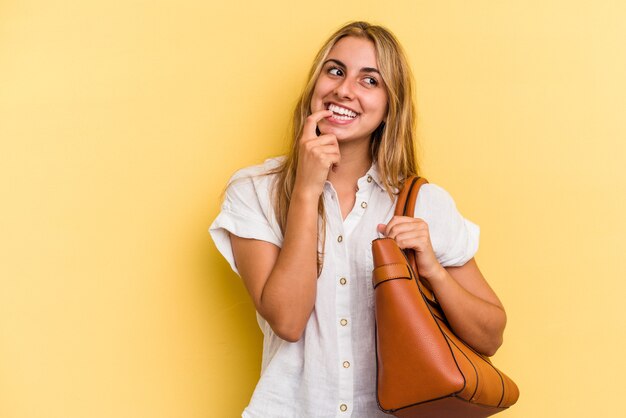 Giovane donna bionda caucasica che indossa una borsa in pelle isolata su sfondo giallo rilassata pensando a qualcosa guardando uno spazio di copia.