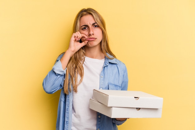 Giovane donna bionda caucasica che tiene le pizze isolate su sfondo giallo con le dita sulle labbra mantenendo un segreto.