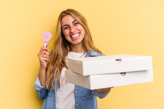 Giovane donna bionda caucasica che tiene pizze e taglierina isolati su sfondo giallo