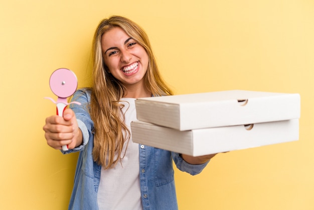 Giovane donna bionda caucasica che tiene pizze e taglierina isolati su sfondo giallo