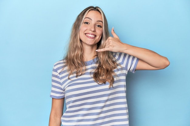 Giovane donna bionda caucasica in studio blu che mostra un gesto di chiamata al telefono cellulare con le dita