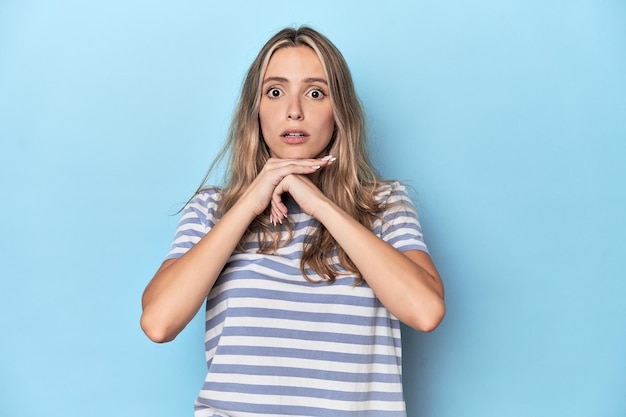 Giovane donna bionda caucasica in studio blu che prega per la fortuna sbalordita e che apre la bocca