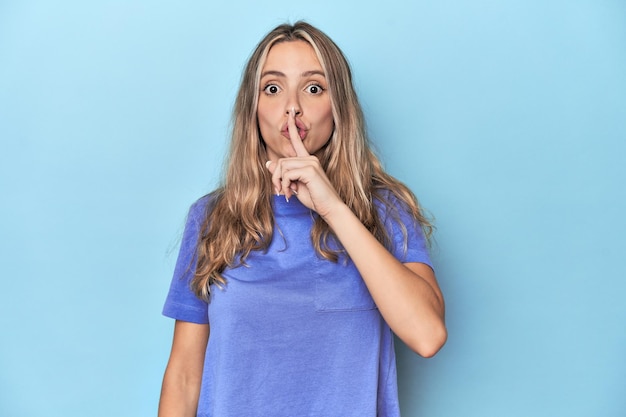 Giovane donna bionda caucasica in studio blu che tiene un segreto o chiede silenzio.