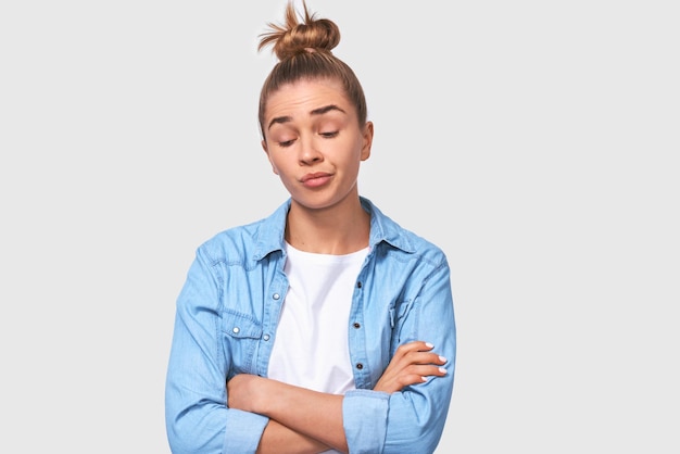 Giovane donna bionda caucasica infelice con le mani incrociate che indossa abiti casual con crocchia per capelli Studentessa piuttosto insoddisfatta che posa sopra il muro bianco dello studio Concetto di persone ed emozioni