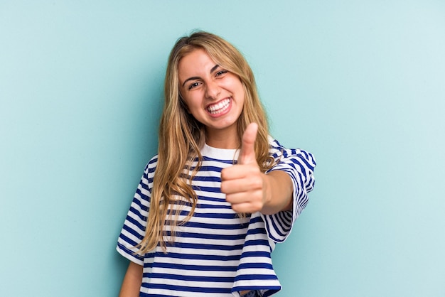 Giovane donna bionda caucasica isolata su sfondo blu sorridente e alzando il pollice