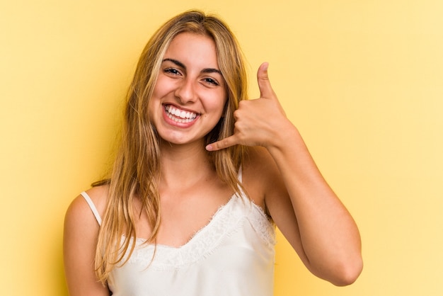 Giovane donna bionda caucasica isolata su sfondo giallo che mostra un gesto di telefonata con le dita.