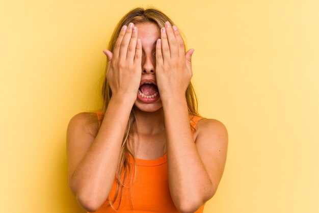 Giovane donna bionda caucasica isolata su sfondo giallo che si diverte a coprire metà del viso con il palmo.