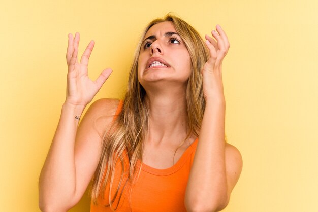 Giovane donna bionda caucasica isolata su sfondo giallo che urla al cielo, alzando lo sguardo, frustrata.