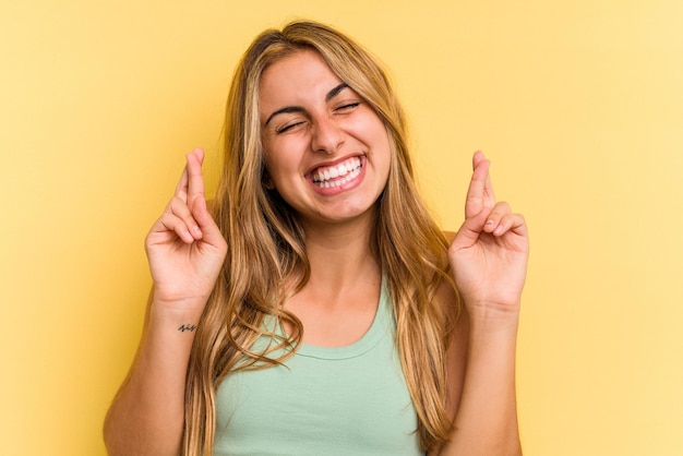 Giovane donna bionda caucasica isolata su sfondo giallo incrociando le dita per avere fortuna